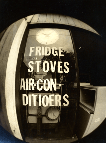 A fisheye black and white photograph of a shop window reading 'FRIDGE STOVES AIR-CON-DITIOERS' [sic]